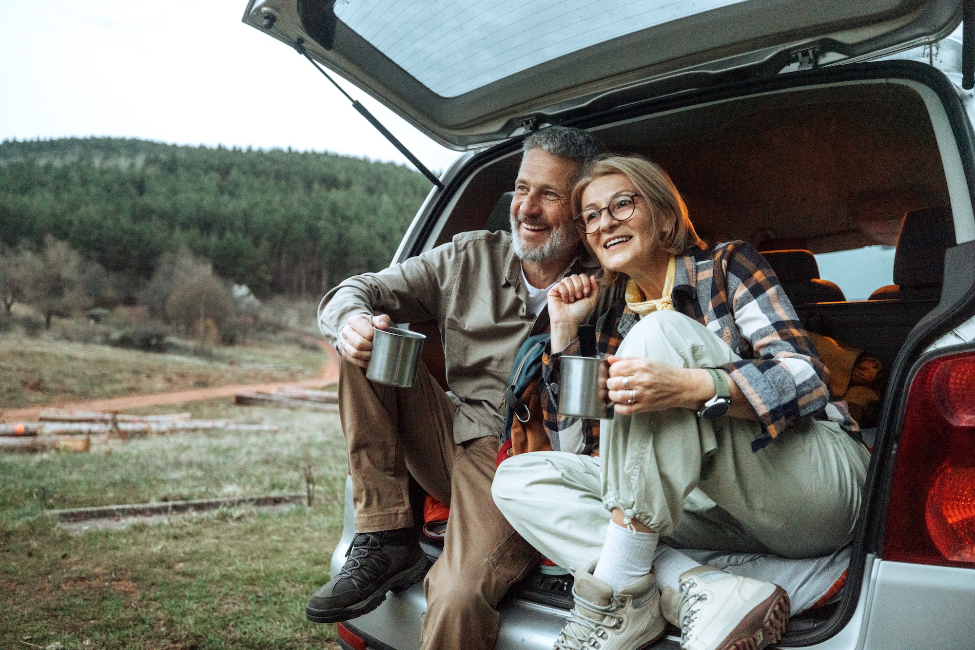 Older couple sitting in an open car trunk, smiling with mugs, forested hillside behind them.