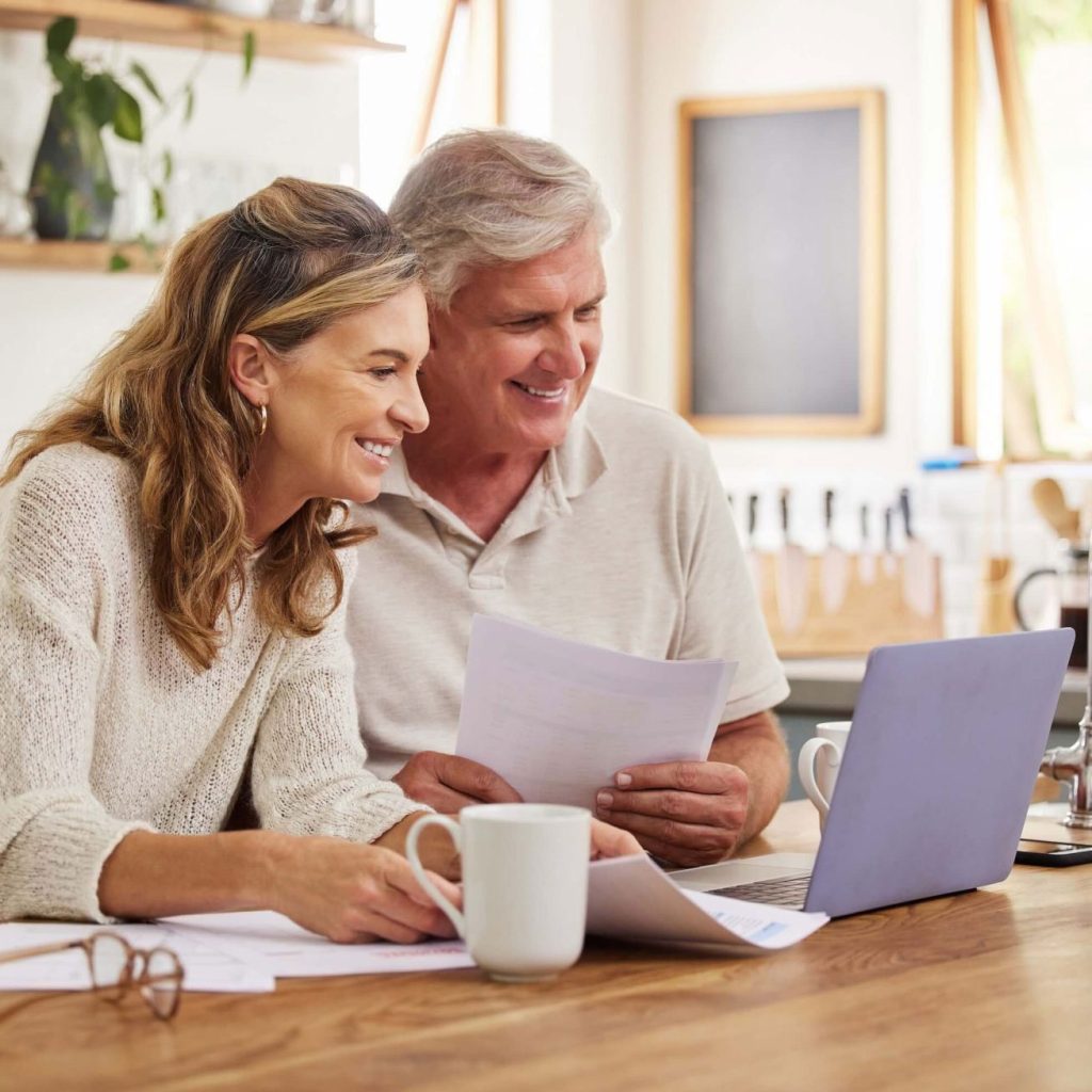 Couple reviewing paper documents at a kitchen table with a laptop and coffee mugs nearby.